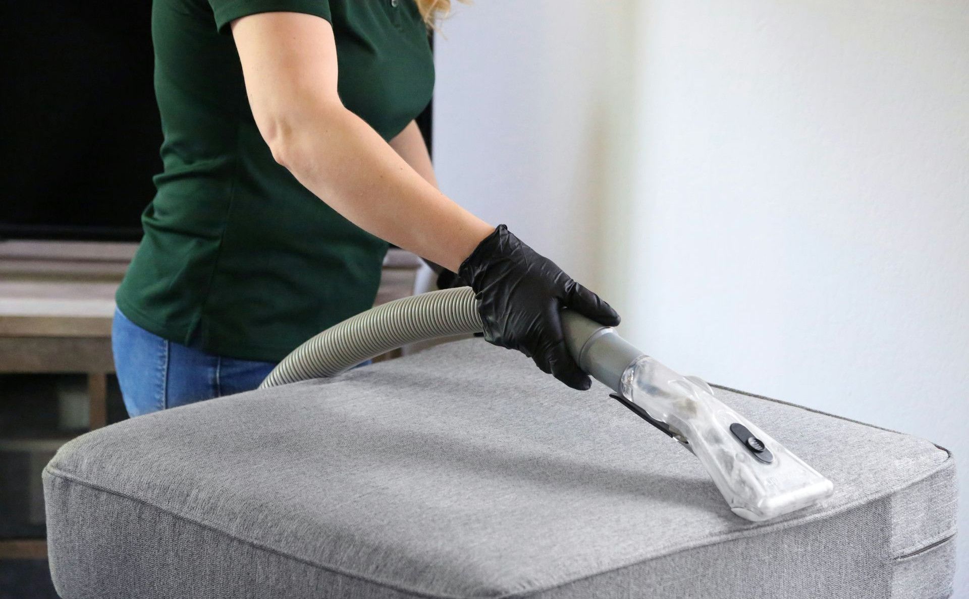 a woman in a green shirt and black gloves vacuuming a gray ottoman